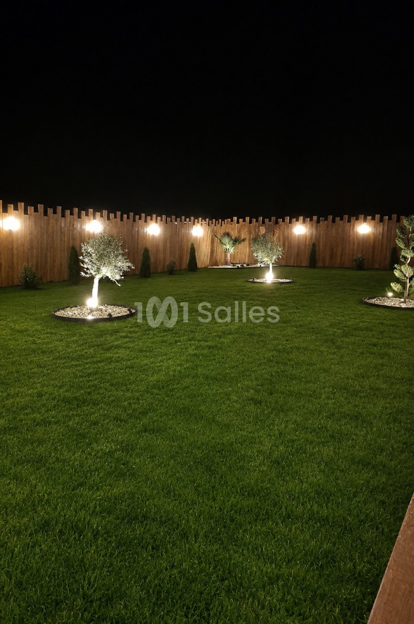 Jardin de nuit avec pelouse verte, clôture bois, arbres éclairés et luminaires suspendus.
