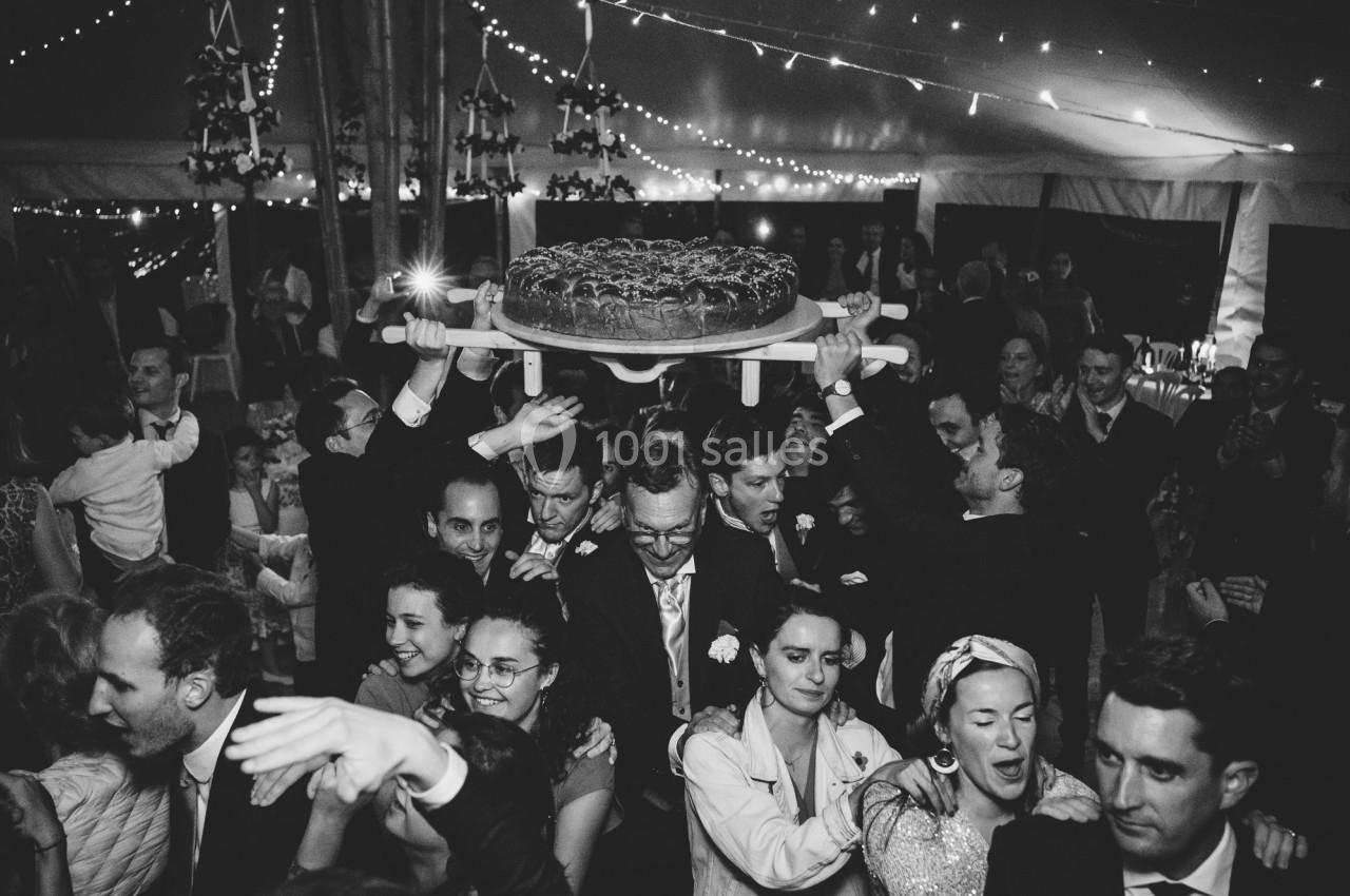 Des invités dansent sous une tente éclairée, portant un plateau avec une grande brioche au-dessus de la foule.