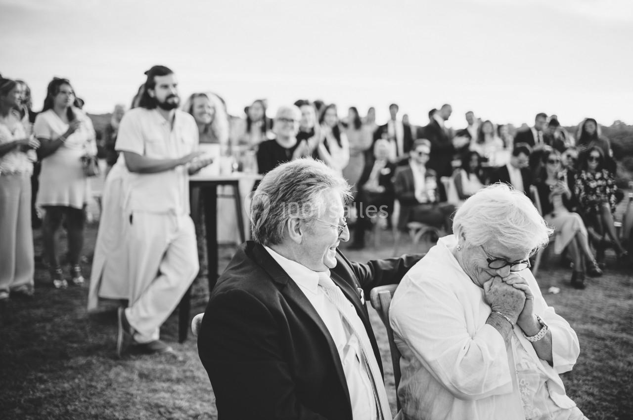 Un couple âgé assis, échangeant un moment tendre, entouré d'invités lors d'un événement en plein air.