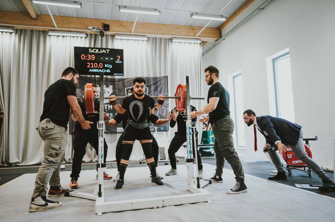 Un homme réalise un squat avec une barre chargée, entouré de plusieurs assistants dans une salle de sport.