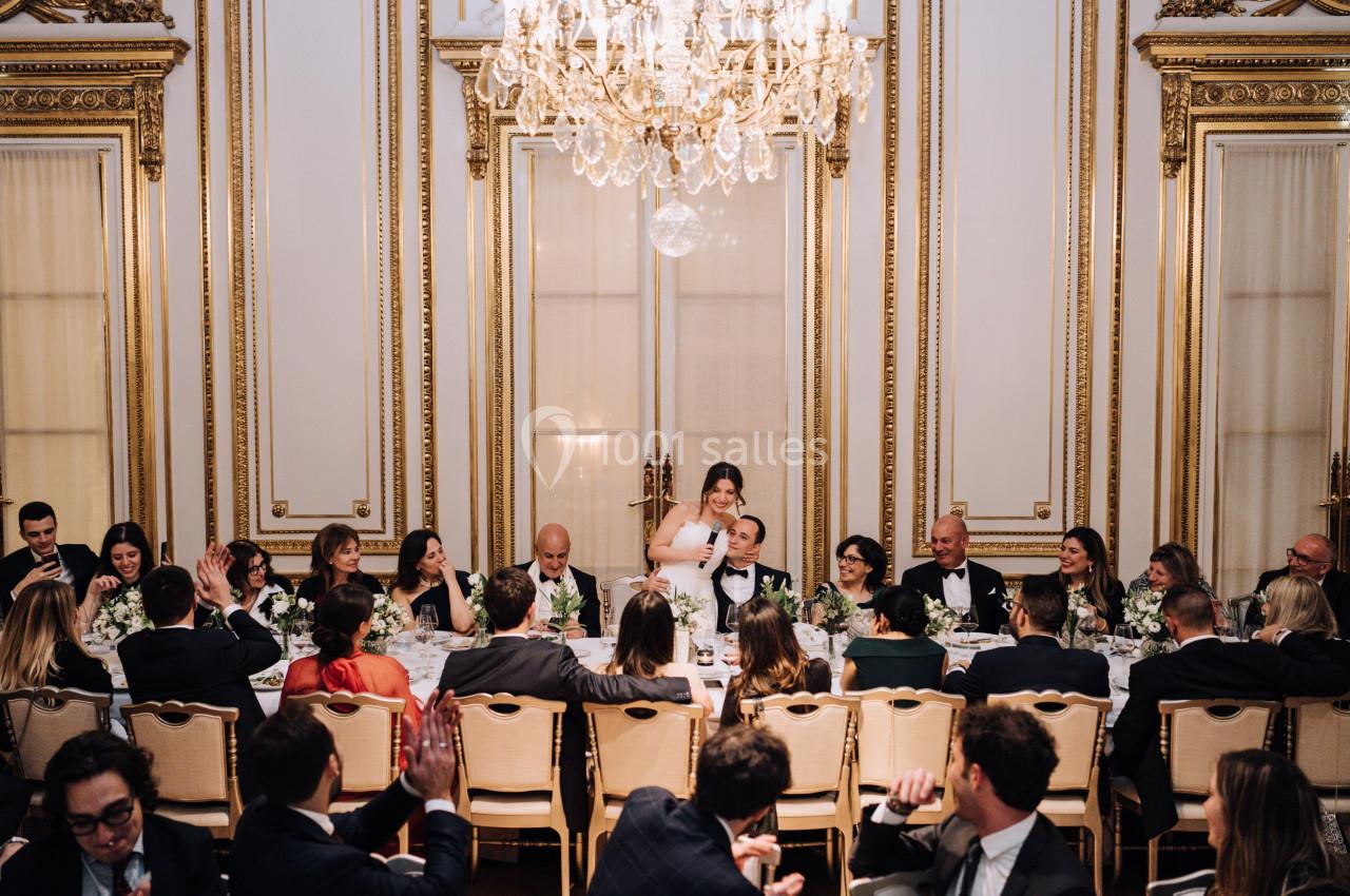 Un dîner formel dans une salle ornée de dorures, avec des invités assis autour d'une longue table décorée de fleurs.