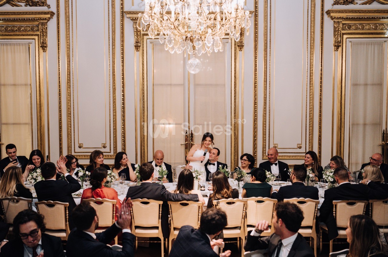 Un dîner formel dans une salle ornée de dorures, avec des invités assis autour d'une longue table décorée de fleurs.