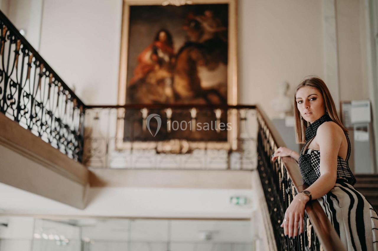 Une femme pose près d'une rampe d'escalier dans un intérieur lumineux avec un grand tableau en arrière-plan.