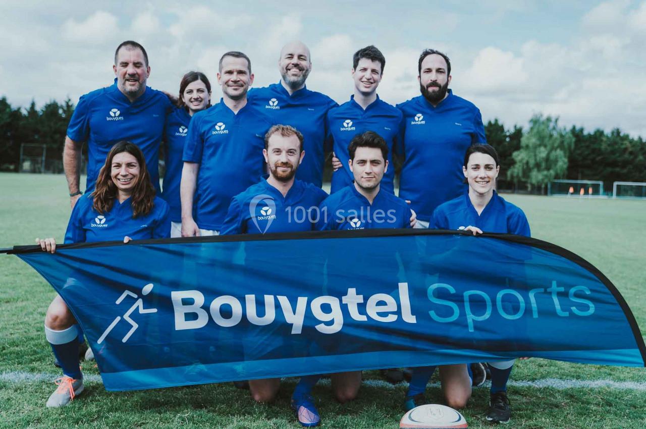 Un groupe de personnes en tenue de sport bleue pose sur un terrain de football avec une bannière ’Bouygtel Sports’.