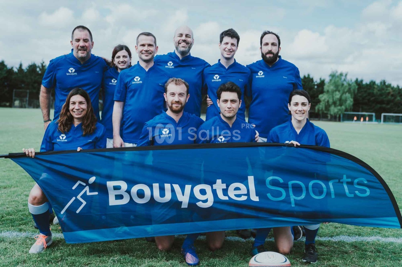 Un groupe de personnes en tenue de sport bleue pose sur un terrain de football avec une bannière ’Bouygtel Sports’.