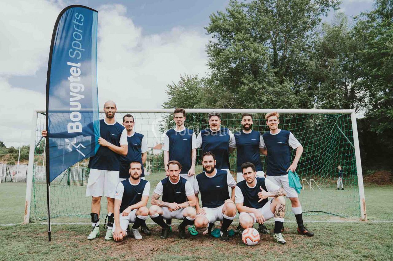 Une équipe de football posant devant un but sur un terrain en plein air, avec un drapeau ’Bouygues Sports’ à gauche.