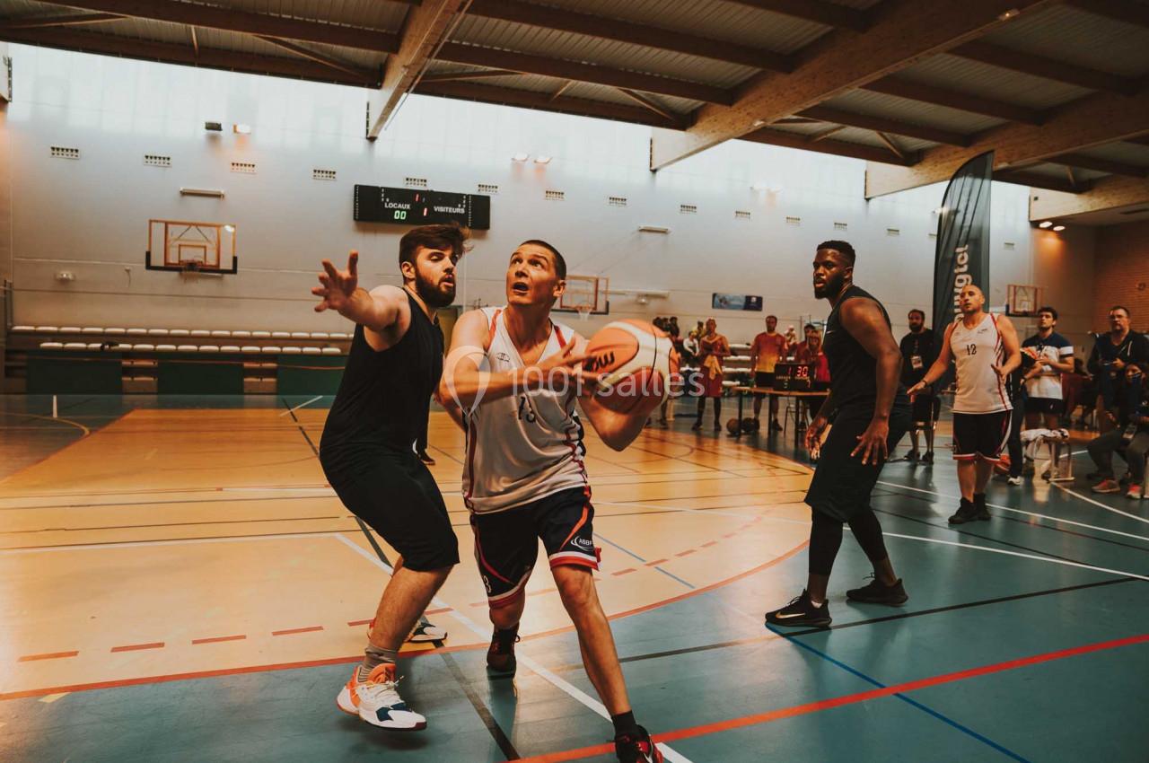 Deux joueurs de basketball en action sur un terrain intérieur, entourés de spectateurs et d'autres joueurs.
