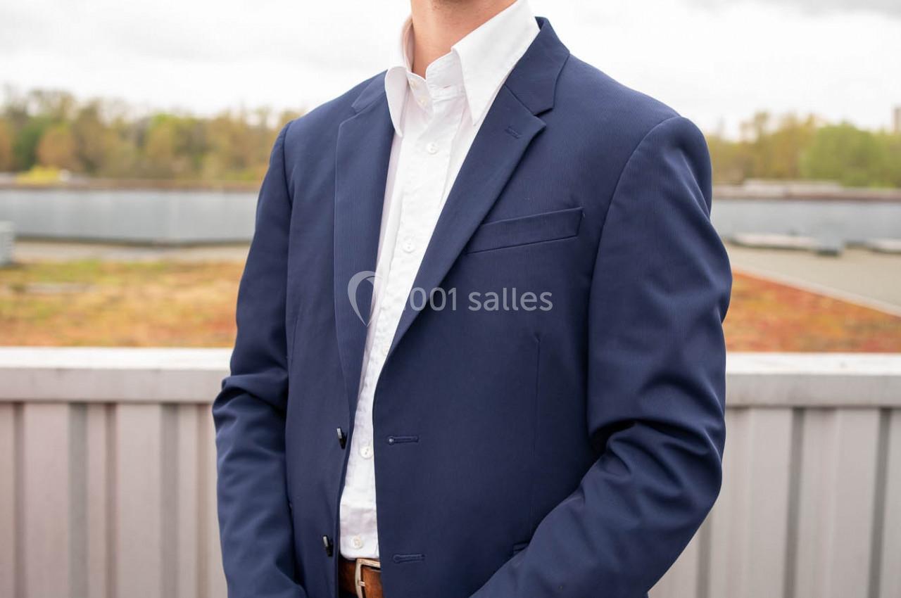 Un homme en costume bleu se tient debout sur une terrasse extérieure par temps nuageux.