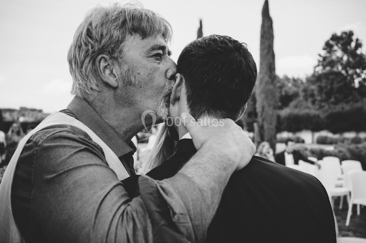 Un homme âgé embrasse affectueusement un homme plus jeune sur la joue dans un cadre extérieur.