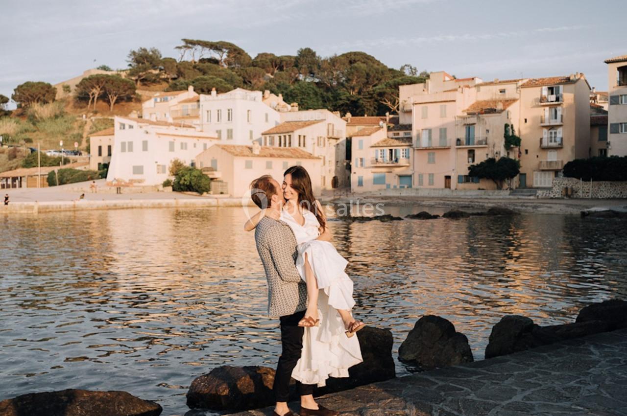 Un couple au bord d'un plan d'eau, l'homme portant la femme dans ses bras devant des bâtiments clairs.