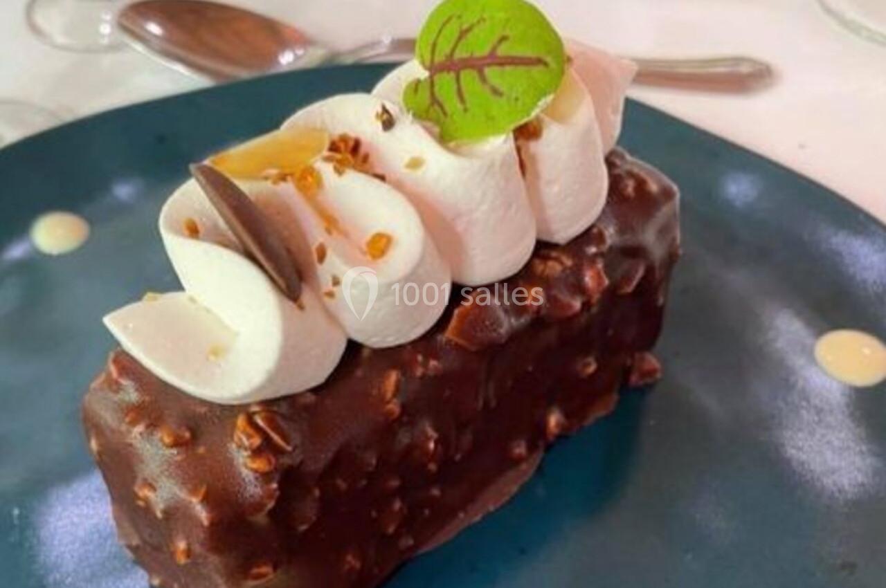 Dessert rectangulaire enrobé de chocolat avec crème fouettée, éclats de noisettes et feuille décorative sur une assiette…