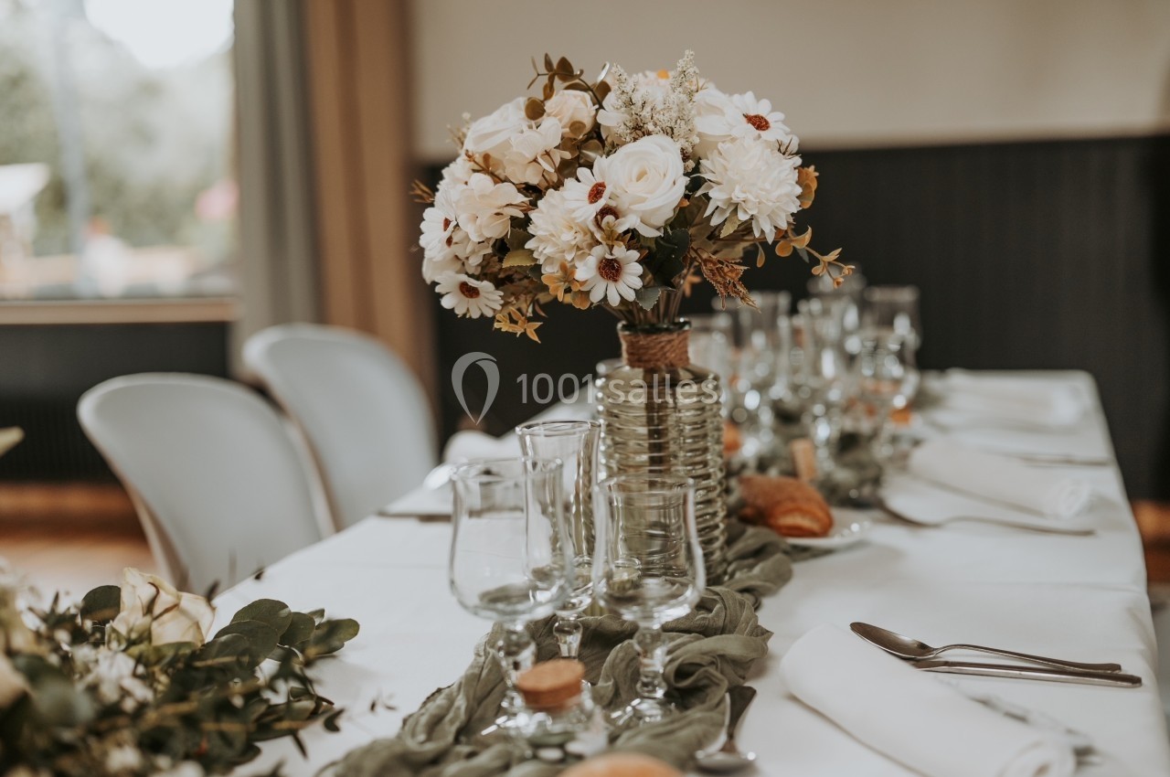 Centre de table floral avec des fleurs blanches et feuillage, disposé sur une table dressée avec vaisselle et serviettes.