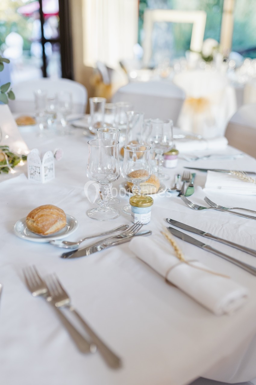 Table dressée avec nappes blanches, couverts, verres, petits pains et décorations pour un repas formel.