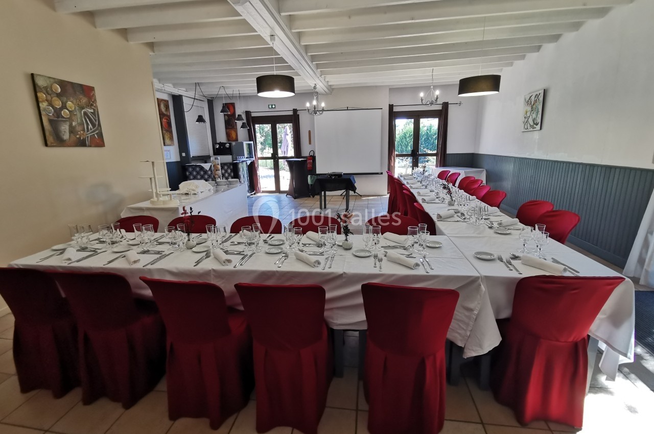 Salle de réception avec deux longues tables dressées, nappes blanches, chaises rouges et lumière naturelle.
