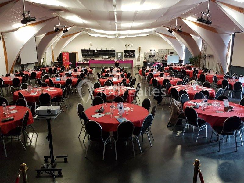 Salle de conférence avec des tables rondes dressées de nappes rouges, chaises noires et matériel audiovisuel installé.