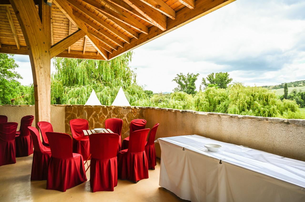 Terrasse couverte avec des chaises rouges autour de tables et une table blanche dressée, vue sur un paysage verdoyant.