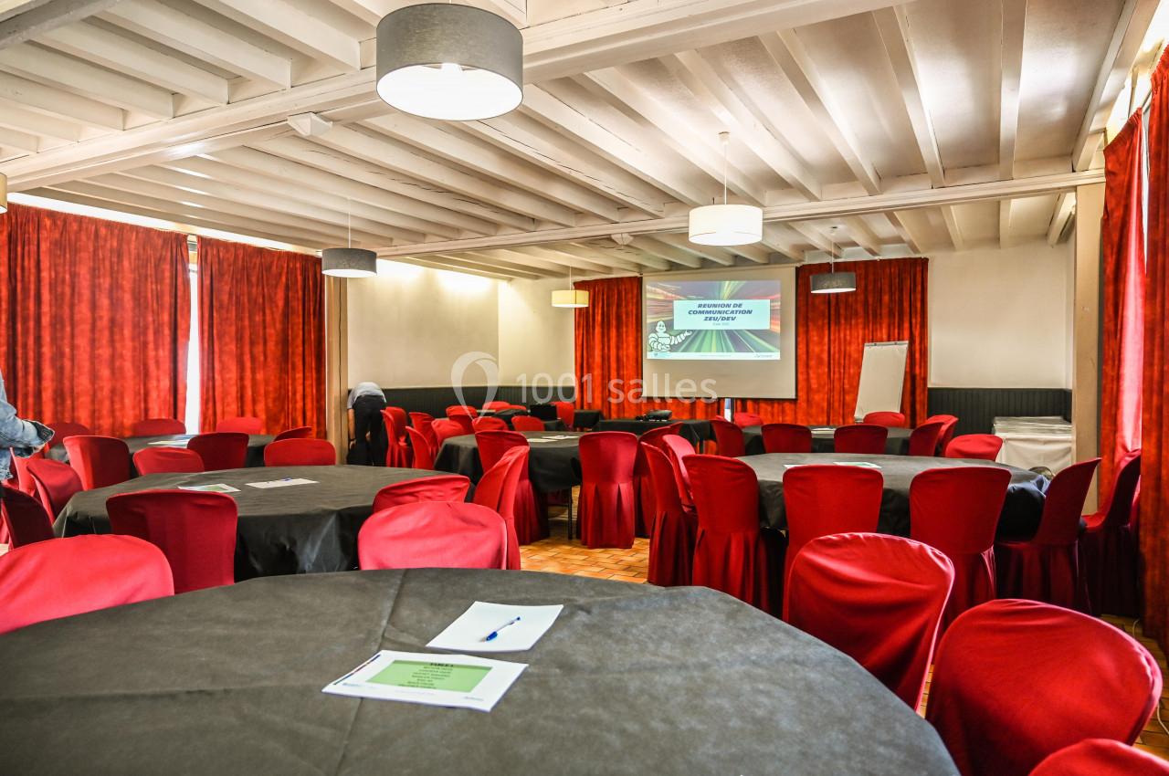 Salle de réunion avec des tables rondes couvertes de nappes noires, chaises rouges et un écran de projection allumé.