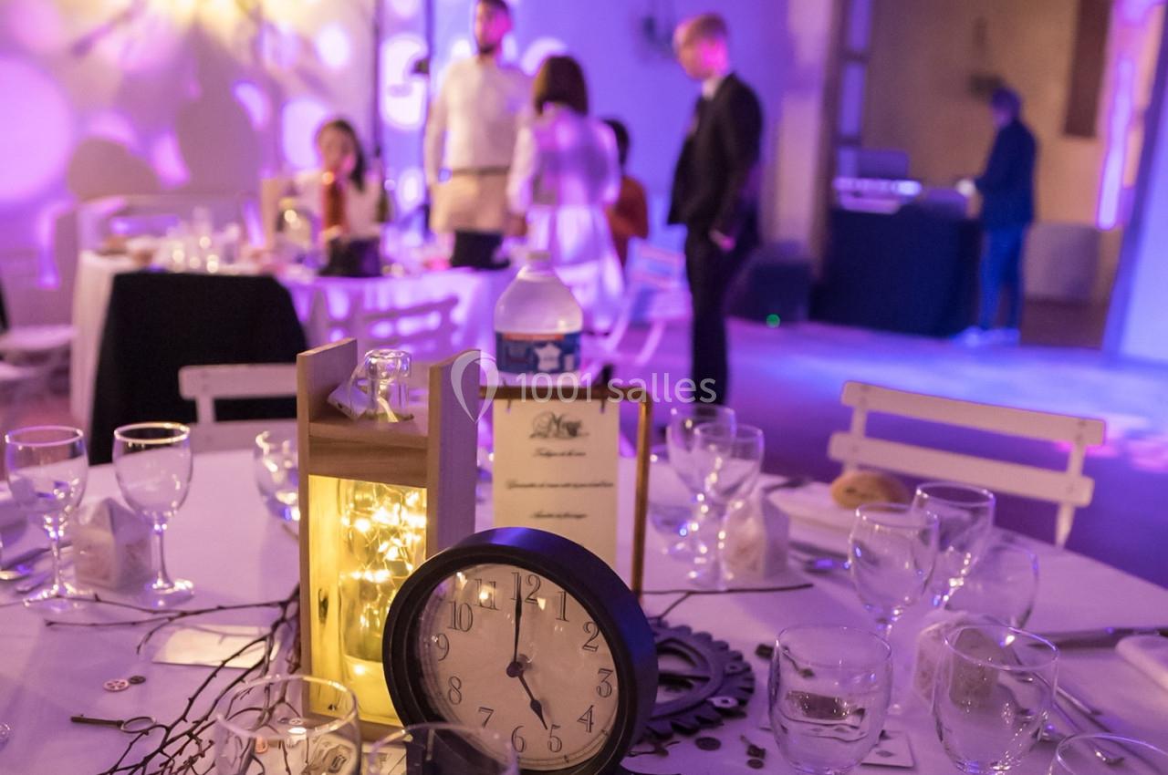 Table décorée avec une horloge, des lumières et des verres, dans une salle de réception avec des invités en arrière-plan.