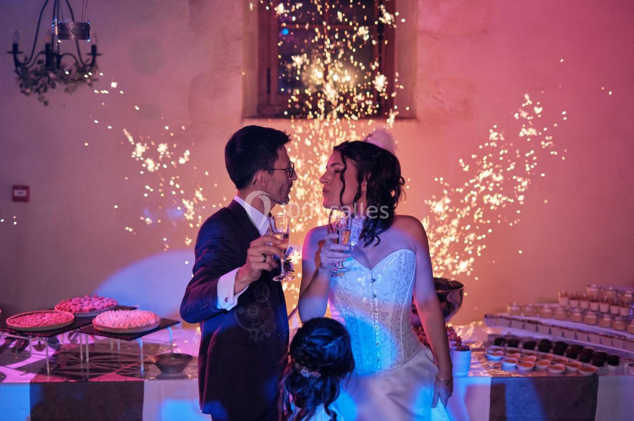 Un couple en tenue de mariage trinque devant une table de desserts, avec des étincelles en arrière-plan.