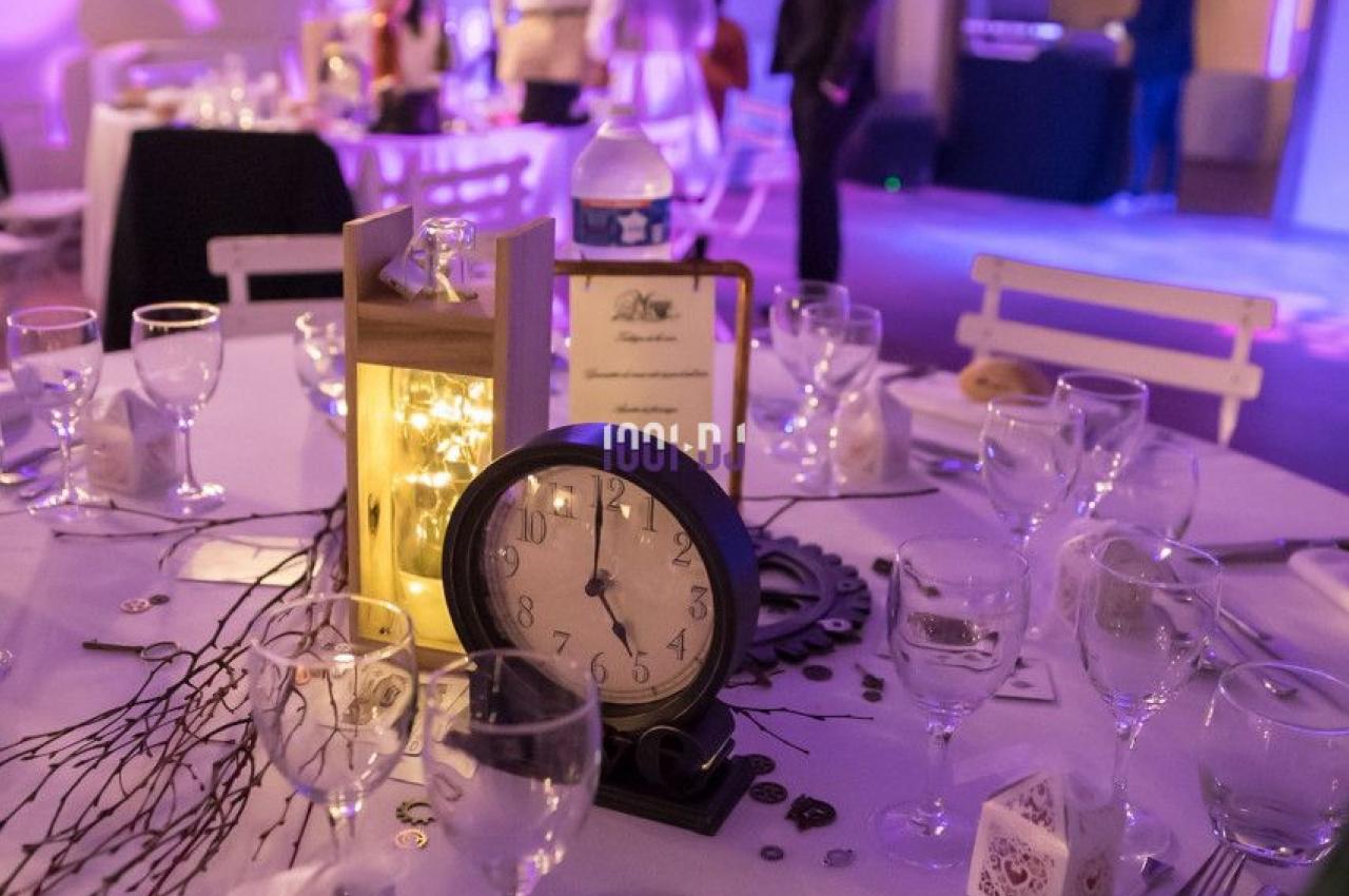 Table décorée pour un événement avec une horloge, des bougies et des branches, dans une salle éclairée en lumière tamisée.