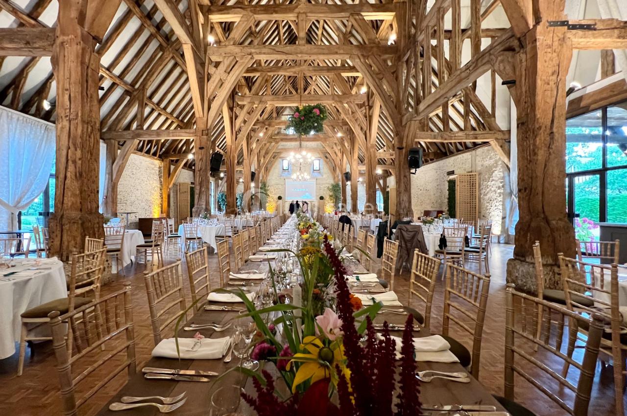 Grande salle de réception avec charpente en bois, tables dressées et décorations florales au centre.