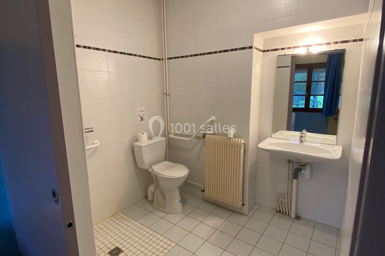 Salle de bain accessible avec toilettes, barre d'appui, lavabo, miroir et carrelage clair.