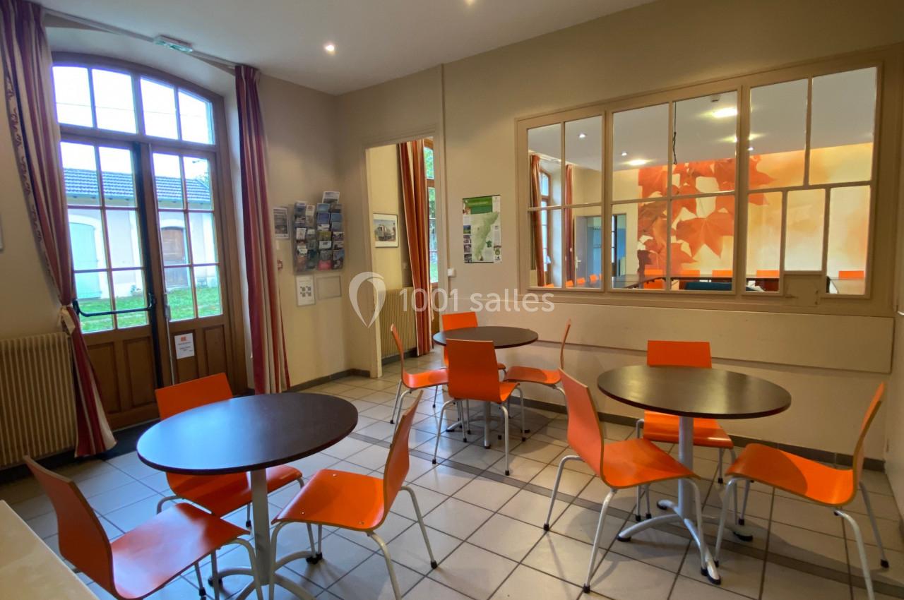 Salle lumineuse avec tables rondes, chaises orange, carrelage clair, grandes fenêtres et panneaux d'affichage au mur.