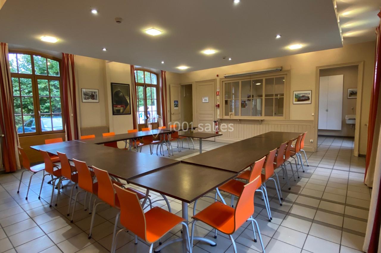 Salle de réunion lumineuse avec tables disposées en U et chaises orange, entourée de grandes fenêtres.