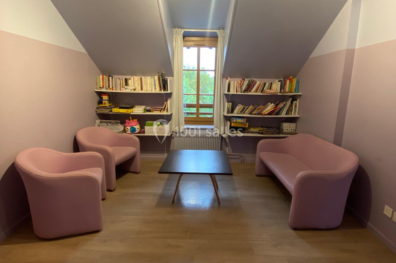 Salon mansardé avec murs violets, deux fauteuils, un canapé rose, une table basse et des étagères remplies de livres.