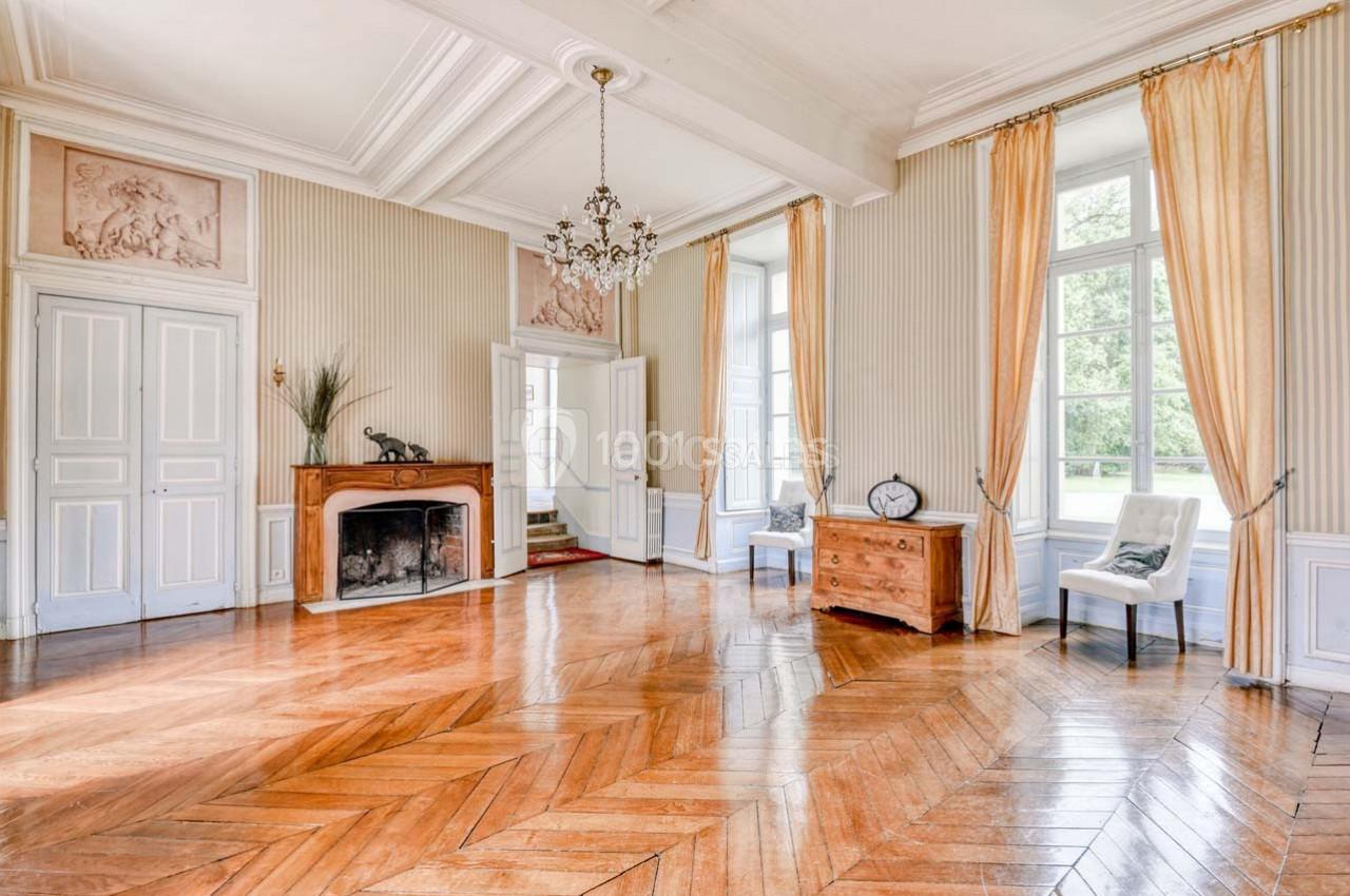 Salle lumineuse avec parquet en bois, cheminée, lustre, grandes fenêtres et mobilier minimaliste.