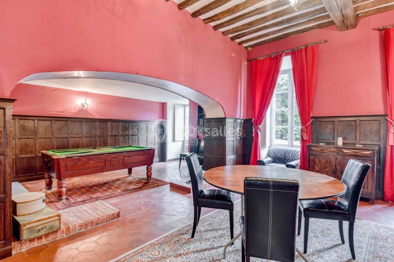 Salle avec murs rouges, billard, table ronde en bois, chaises noires, poutres apparentes et grandes fenêtres avec rideaux.