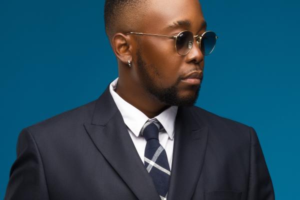 Homme portant un costume sombre, une cravate à motifs et des lunettes rondes, posé sur un fond bleu uni.