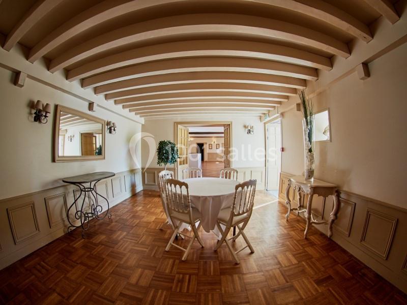 Salle lumineuse avec parquet, table ronde couverte d'une nappe blanche, chaises en bois et poutres apparentes au plafond.