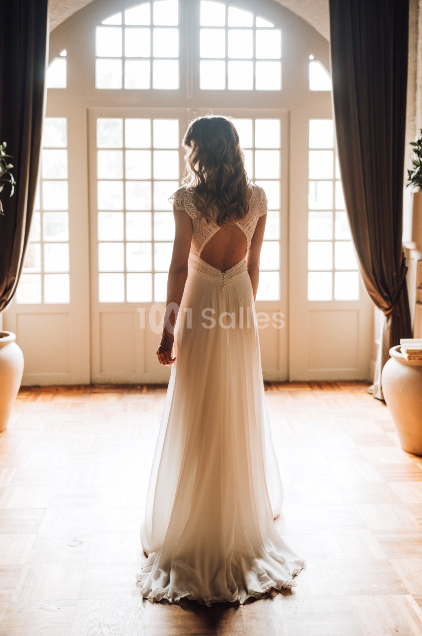 Femme en robe de mariée blanche, vue de dos, devant une grande baie vitrée avec rideaux sombres.