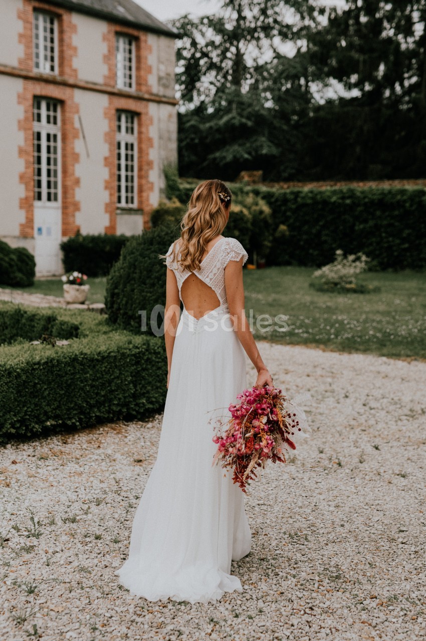 Une femme en robe de mariée blanche tient un bouquet de fleurs roses dans un jardin devant un bâtiment ancien.