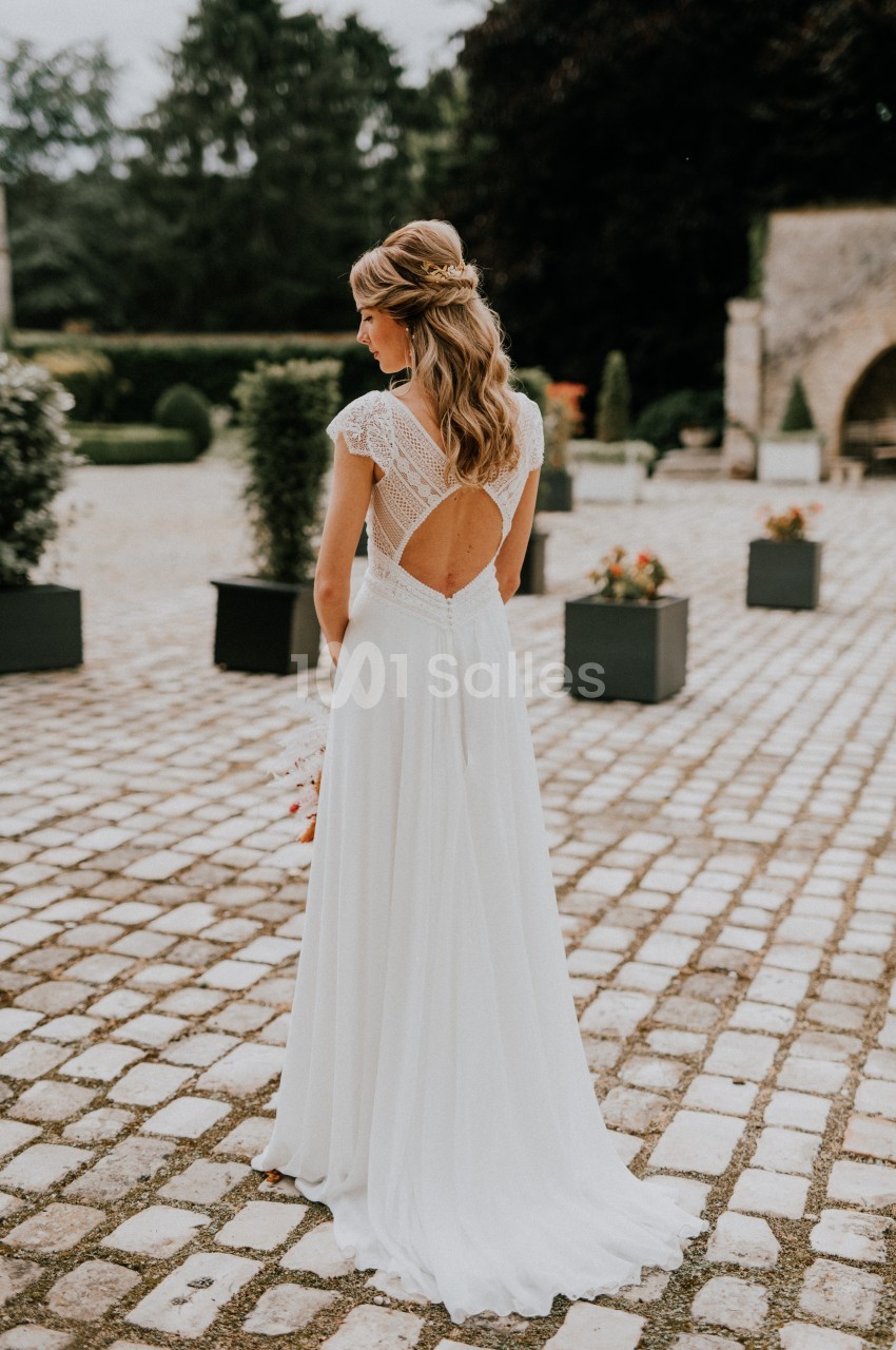Femme en robe de mariée blanche vue de dos, debout sur une allée pavée entourée de verdure et de pots de fleurs.