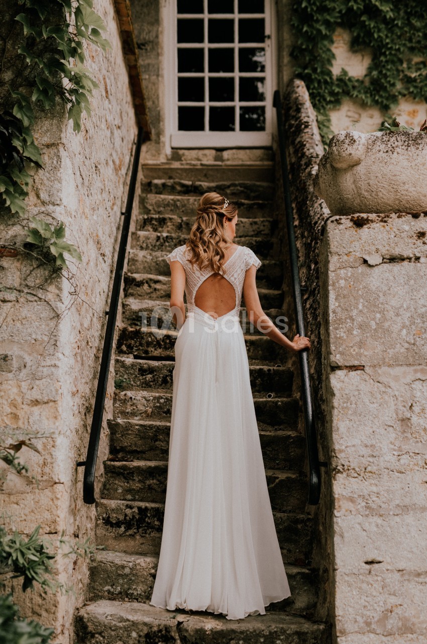 Femme en robe de mariée blanche montant un escalier en pierre, dos tourné, entourée de murs anciens et de végétation.