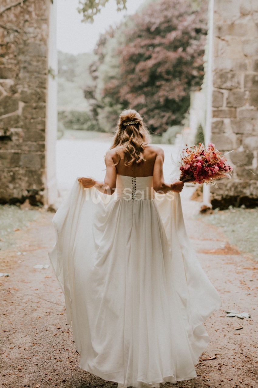 Une femme en robe de mariée blanche tient un bouquet coloré en marchant dans une allée bordée de pierres.