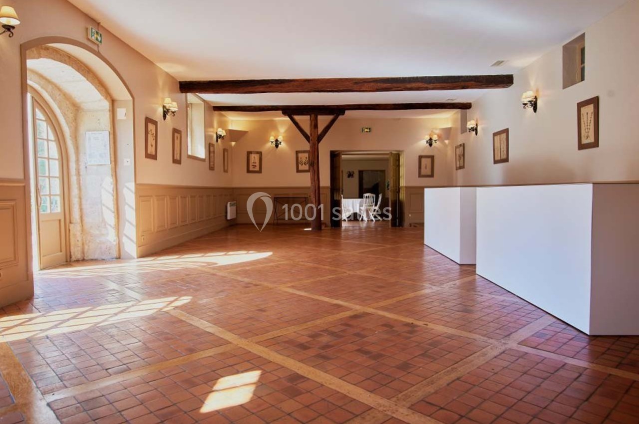 Salle lumineuse avec sol en carrelage, poutres apparentes, murs beige clair et grandes fenêtres laissant entrer la lumière.