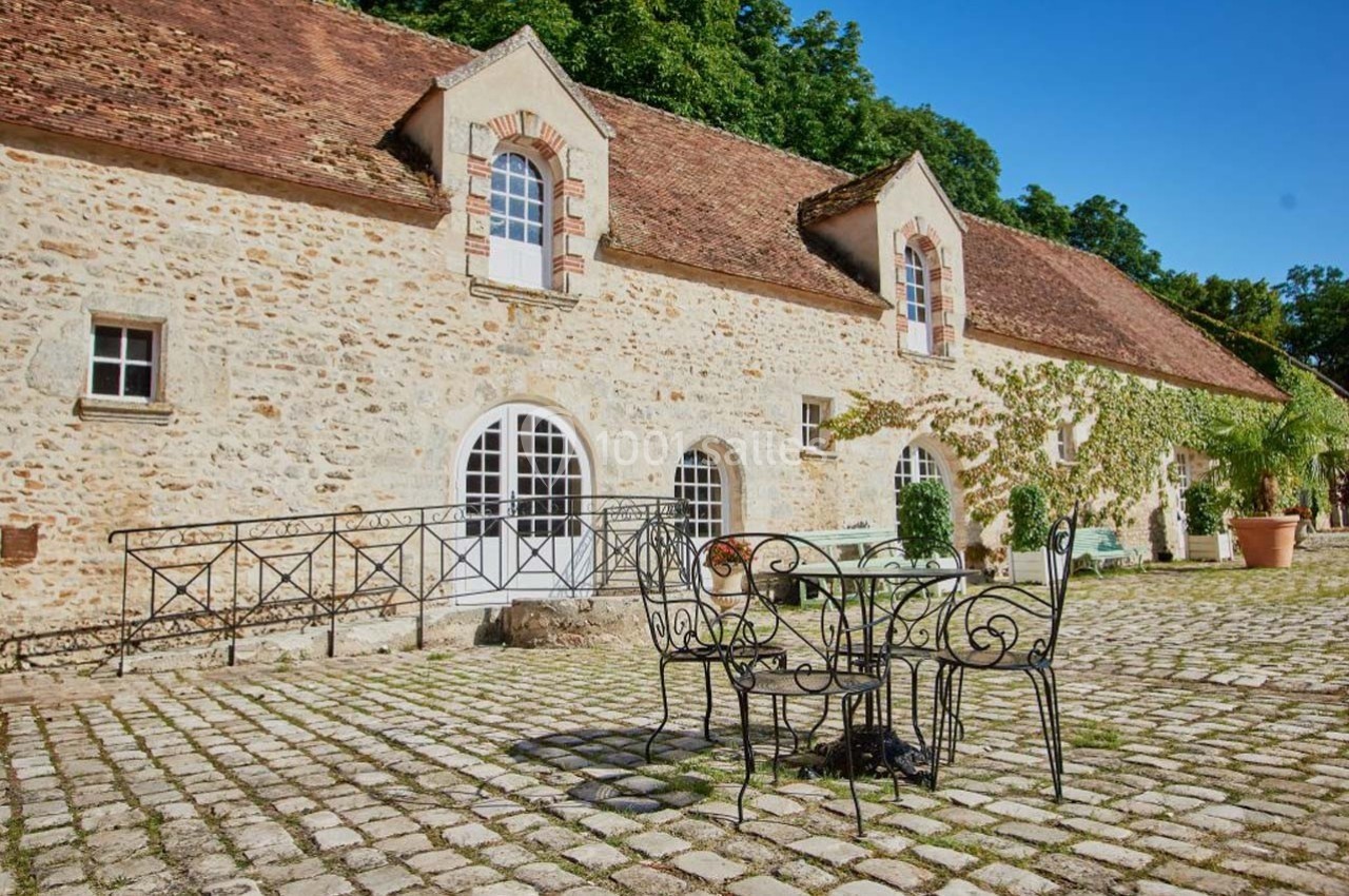 Cour pavée avec table en fer forgé devant un bâtiment en pierre aux toits en tuiles, entouré de verdure.