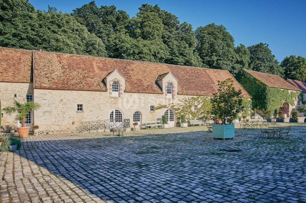 Cour pavée avec un bâtiment en pierre, toit en tuiles, fenêtres cintrées et végétation environnante.
