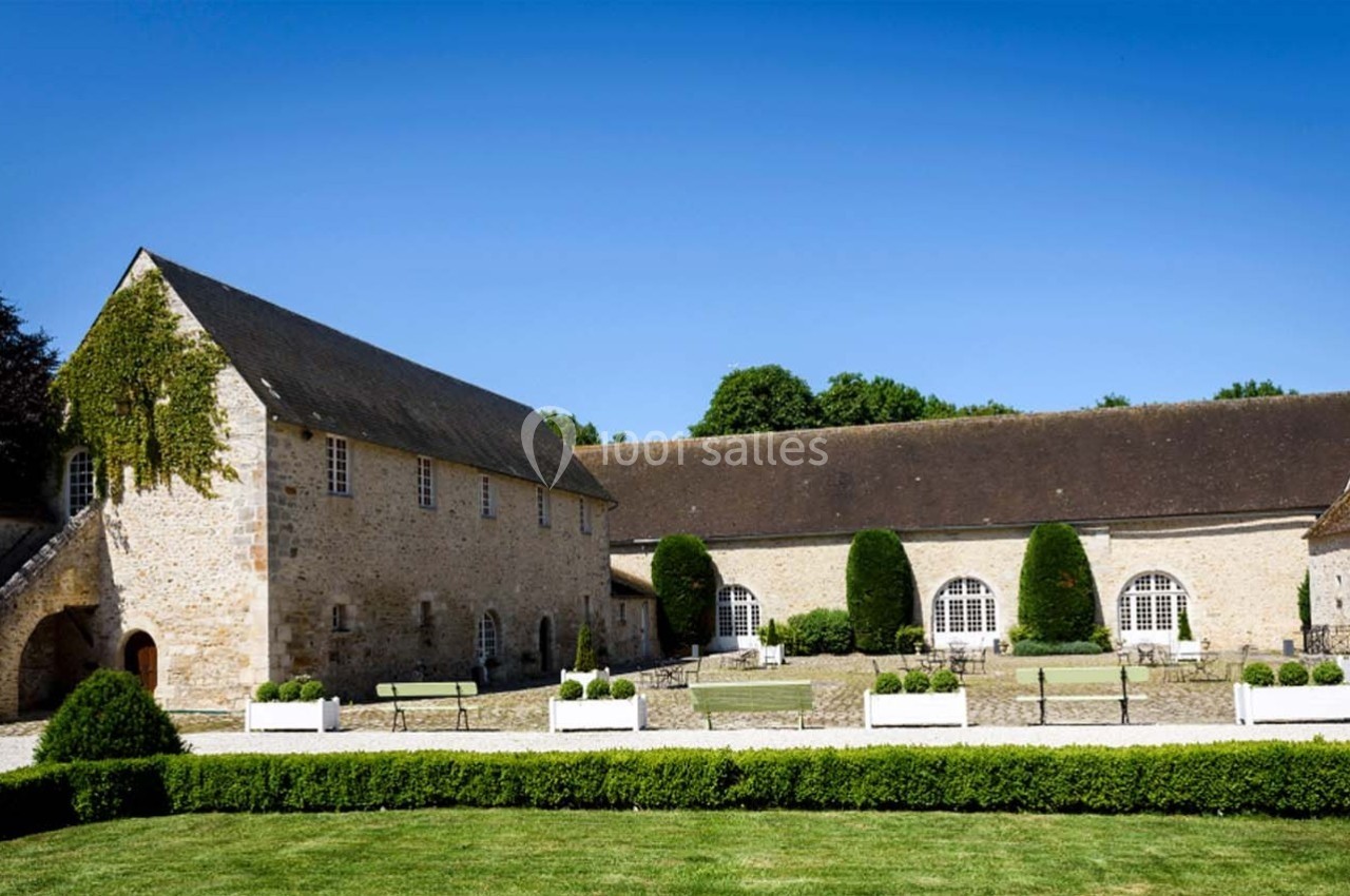 Bâtiments en pierre avec toits en ardoise entourant une cour pavée, bordée de haies et de bancs.