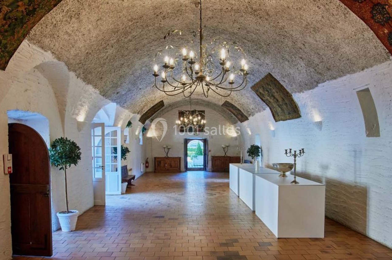 Salle voûtée en pierre blanche avec lustres, tapisseries murales, mobilier minimaliste et lumière naturelle.