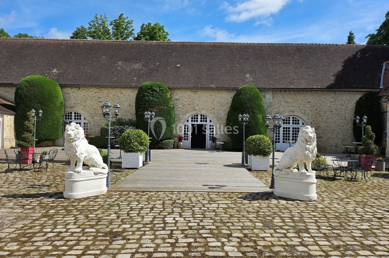 Cour pavée avec deux statues de lions en pierre encadrant une entrée voûtée d'un bâtiment en pierre, entouré de verdure.