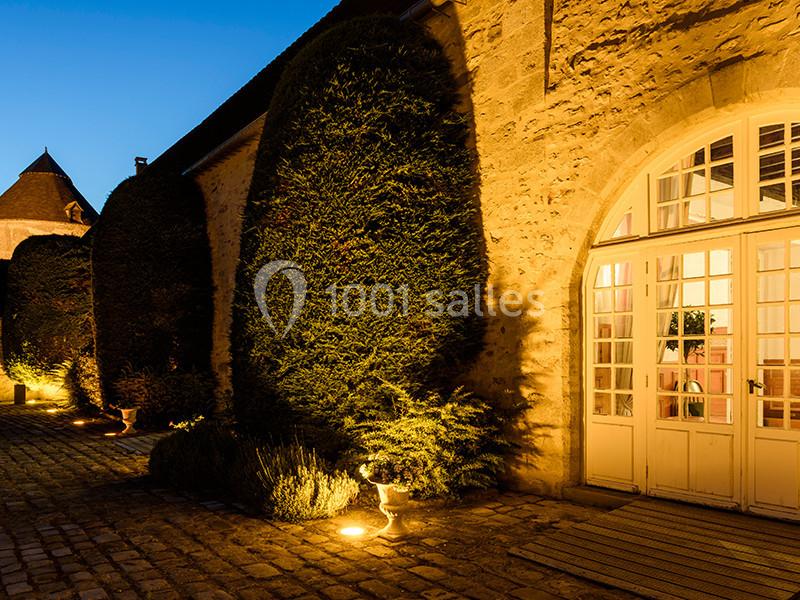 Façade en pierre éclairée de nuit avec grandes fenêtres cintrées et haies taillées dans une cour pavée.