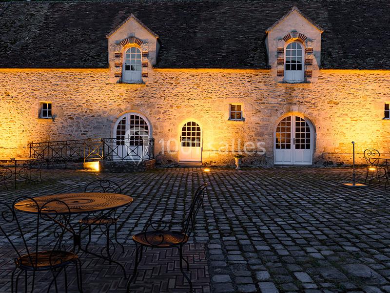 Façade en pierre éclairée de nuit avec des portes et fenêtres cintrées, tables et chaises en fer forgé sur une cour pavée.