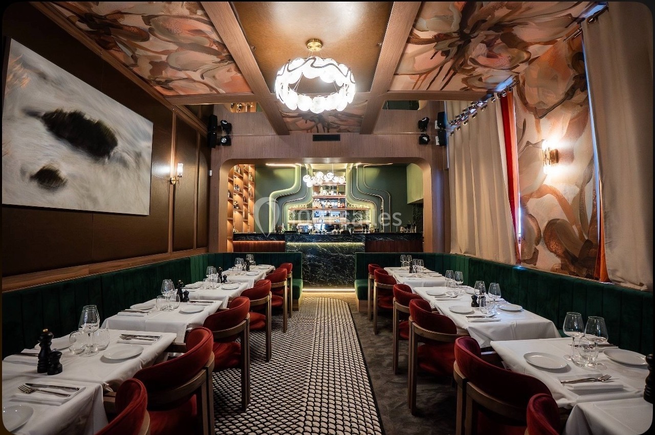 Salle de restaurant élégante avec tables dressées, chaises rouges, éclairage chaleureux et bar en arrière-plan.