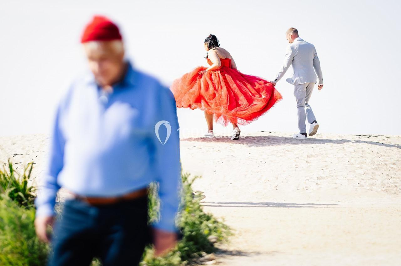 Un couple en tenue élégante marche sur un chemin sablonneux, une femme en robe rouge et un homme en costume gris.