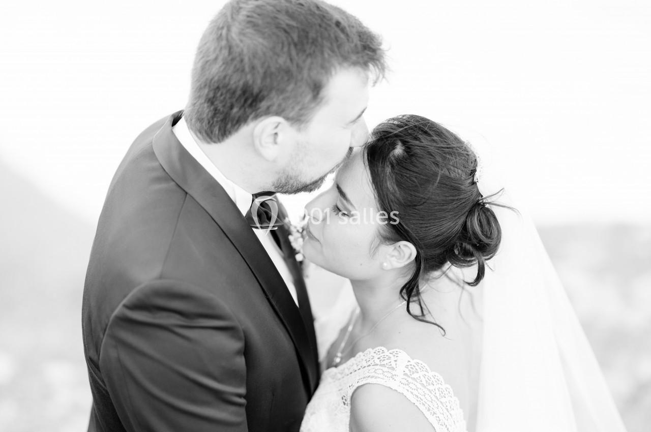Un homme en costume embrasse tendrement une femme en robe de mariée sur le front, en noir et blanc.
