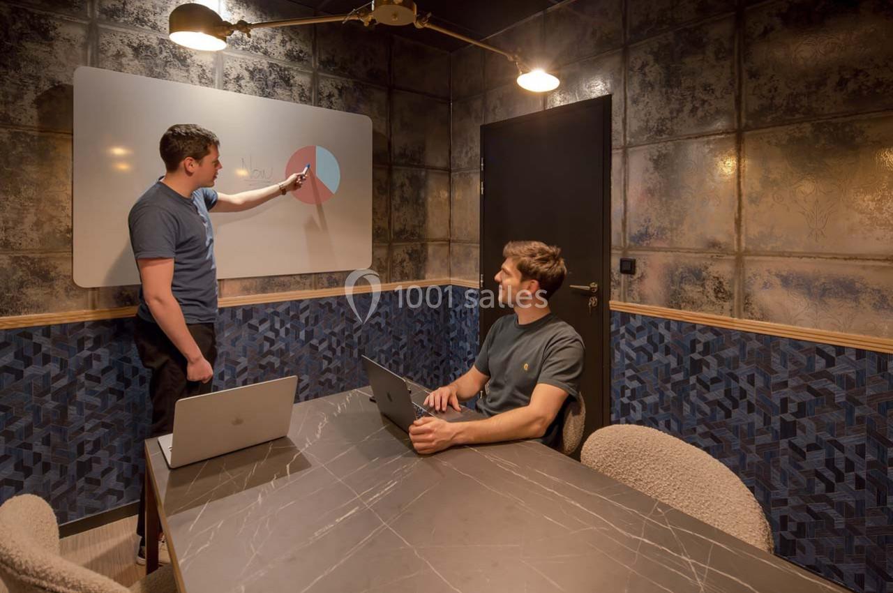 Deux hommes discutent devant un tableau blanc avec un graphique circulaire, dans une salle de réunion moderne.